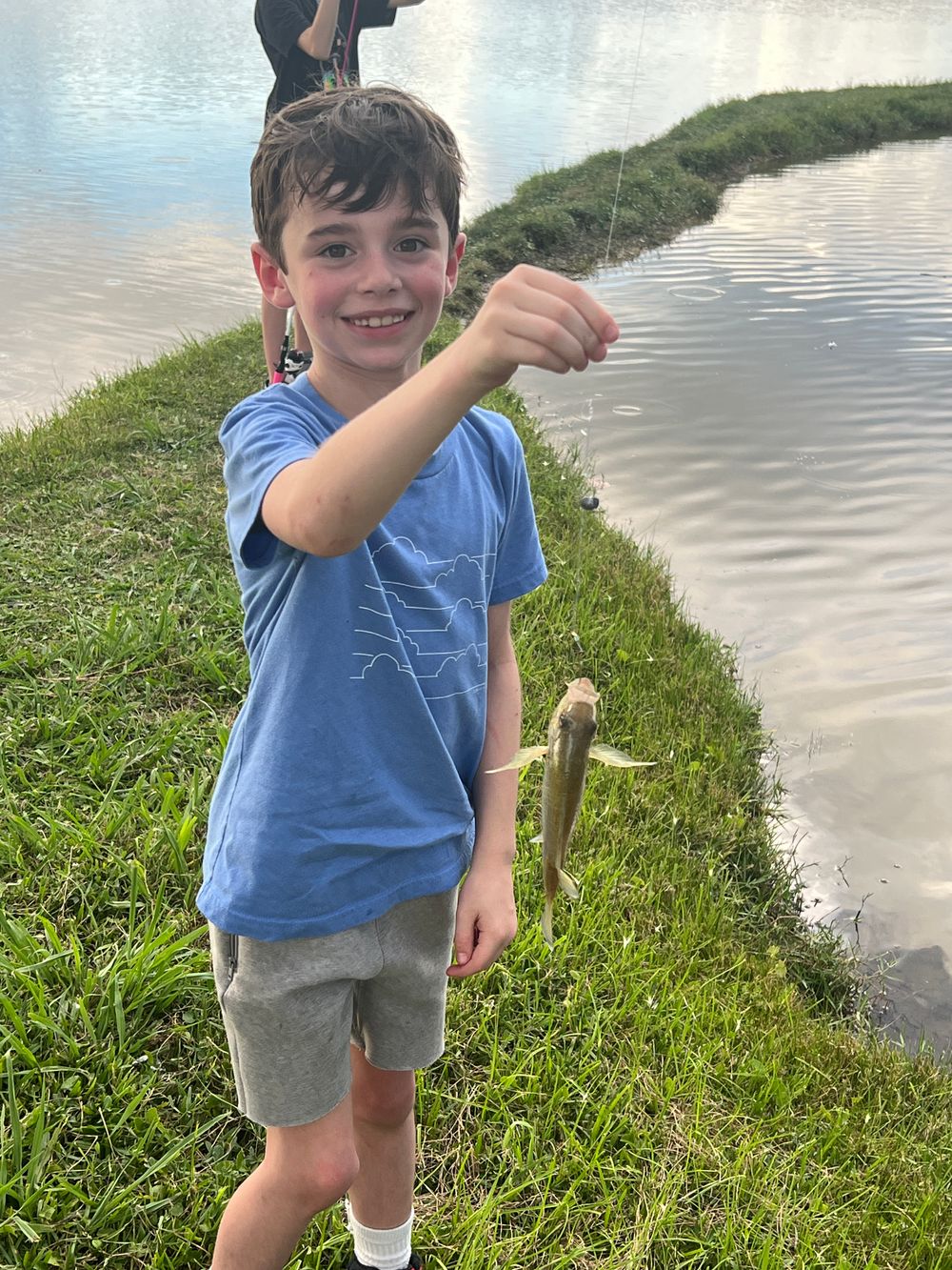 A young gentleman standing on a strip of land that extends out into a lake. He is holdign a small fish on a line.