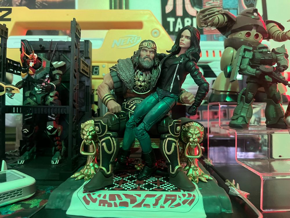 An action figure of a bearded Conan sitting on his throne. Jessica Jones, wearing jeans and a leather jacket, is sitting on his lap.