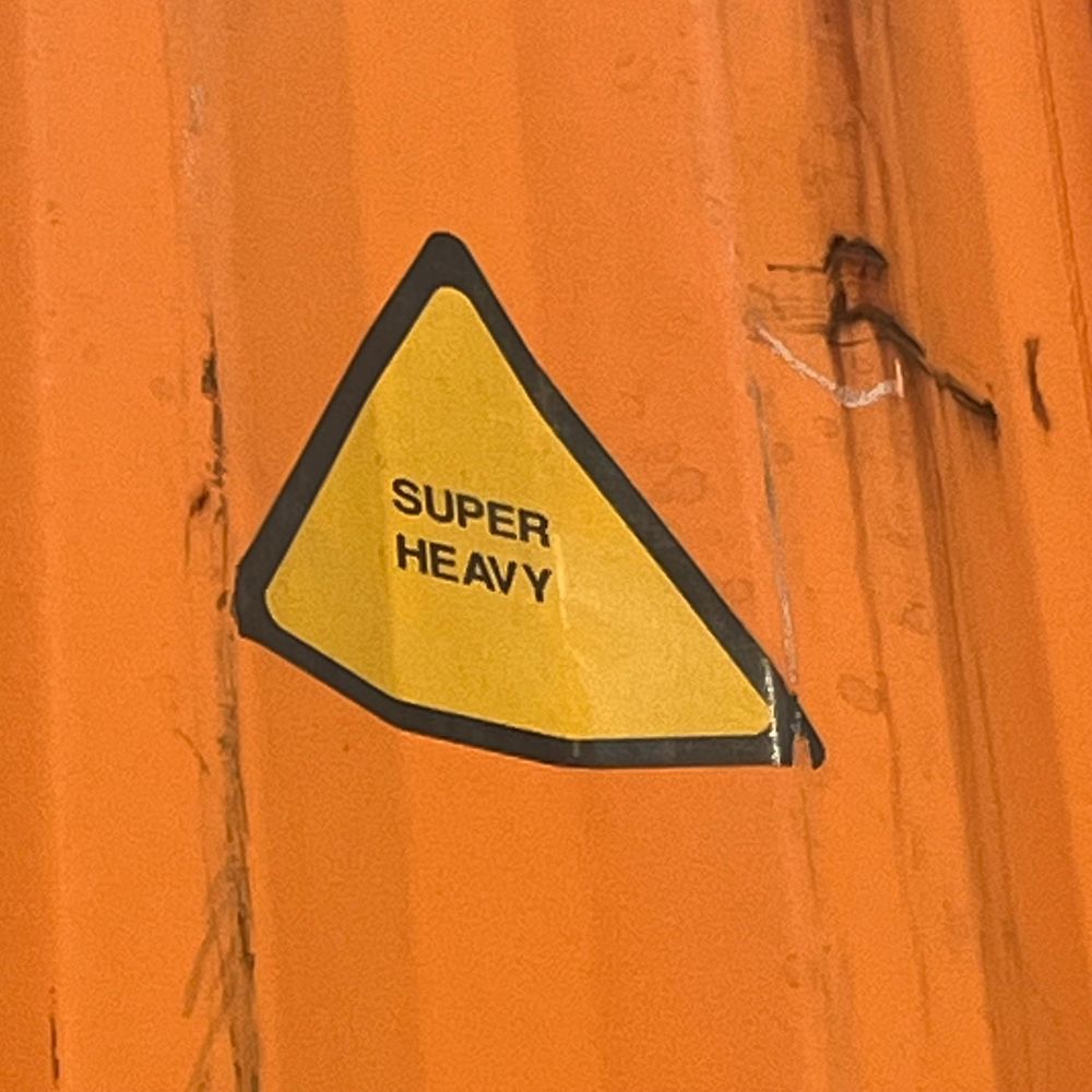 A bright orange shipping container with a yellow triangular sticker that says SUPER HEAVY