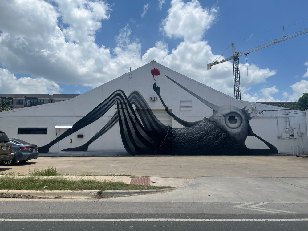 A mural of a weird hummingbird man holding a flower aloft. It is painted on the side of a warehouse.