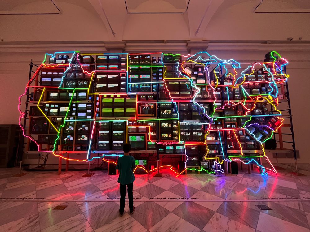 Desmond standing in front of a large neon map of the US with many flickering televisions showing scenes of each state 