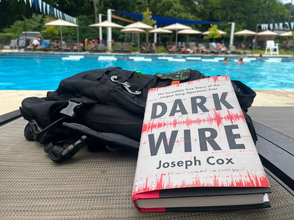 A poolside scene featuring a backpack and a copy of Dark Wire