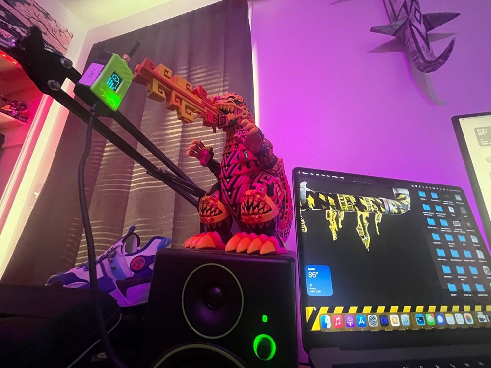 A Godzilla figure standing on a desktop speaker. The Godzilla is decorated with pink and orange Aztec inspired designs. There is a laptop in view and in the background there is a sneaker with the Thundercats logo on it.