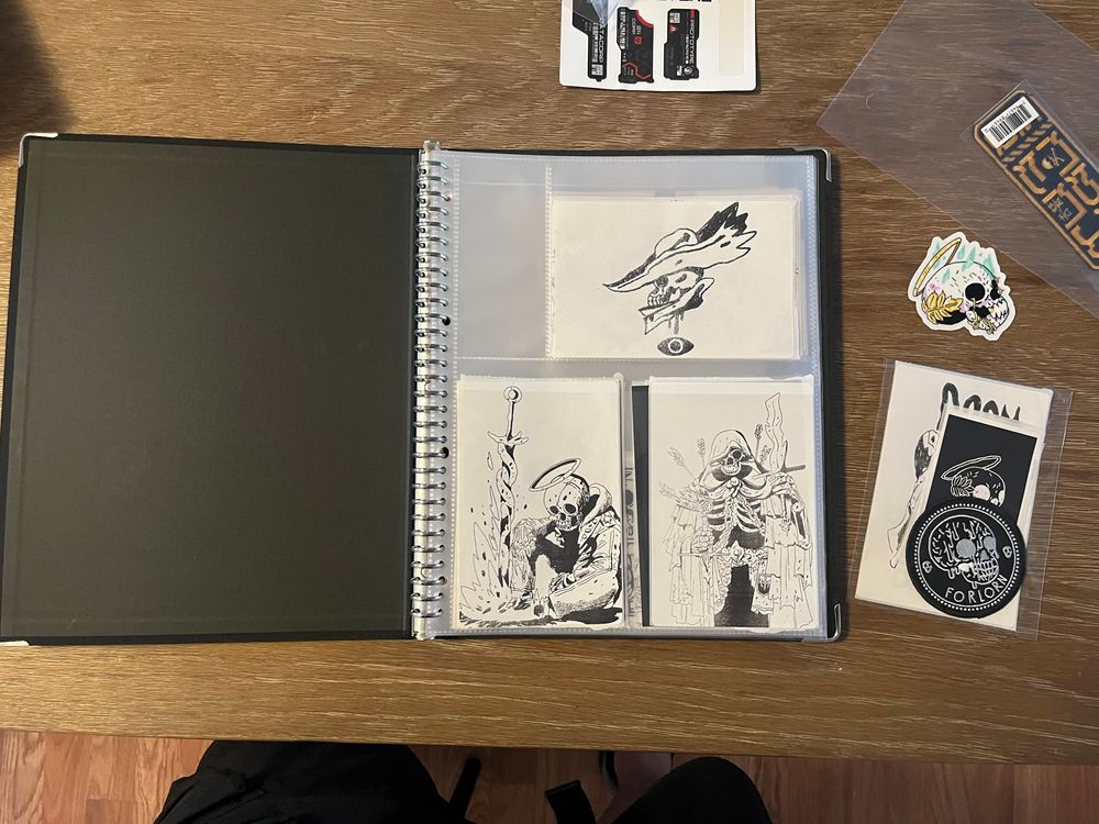 a black binder open on a table. inside is a clear plastic page holding 3 black and white stickers with pictures of skeletons.